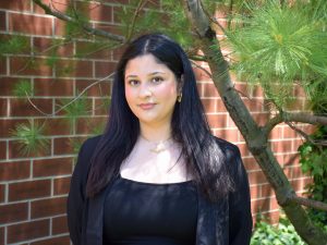 A young woman poses for a photo in front of a brick wall beside a tree.