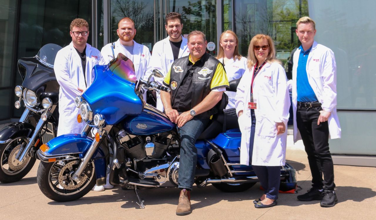 A large group of people pose with two motorcycles outside a glass building. Some of the people are wearing white lab coats.