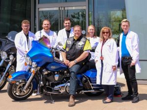 A large group of people pose with two motorcycles outside a glass building. Some of the people are wearing white lab coats.