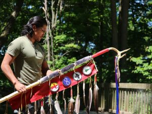 A young First Nations man holds a wooden staff decorated with four beaded emblems and several eagle feathers.