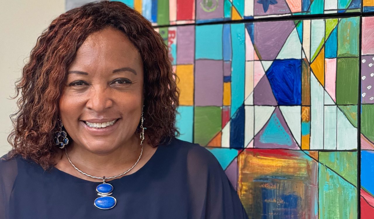 Brock Professor Dolana Mogadime is shown from the shoulders up. She is wearing a deep blue dress with earrings standing in front of a multicoloured abstract painting.