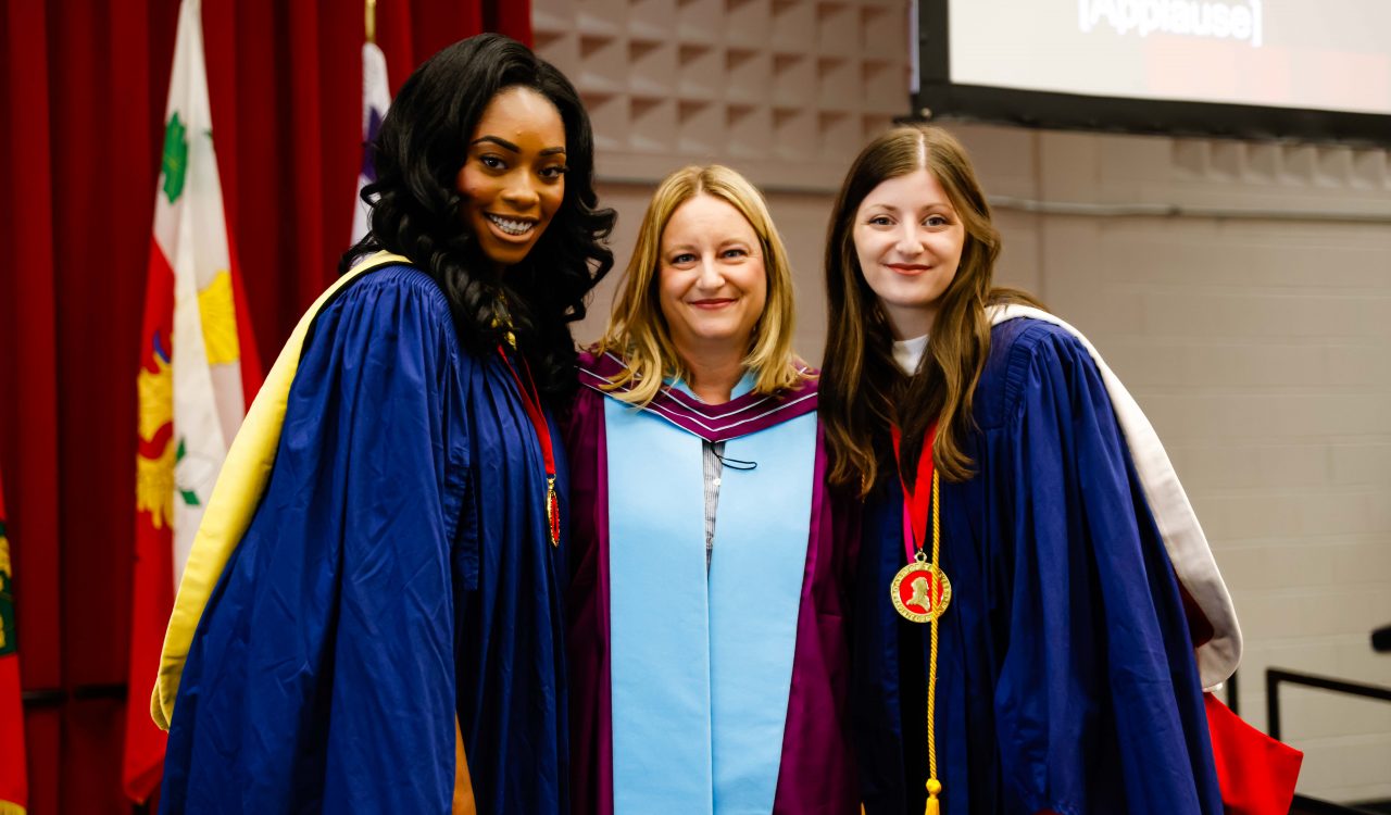 Three women in academic robes pose for a photo on stage during a university graduation ceremony.