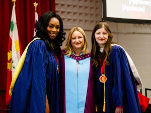 Three women in academic robes pose for a photo on stage during a university graduation ceremony.