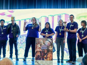 A group of Brock students wearing purple t-shirts stand on a stage in front of a brightly lit digital background. They are wearing medals and smiling at the crowd, and one person is holding a trophy.