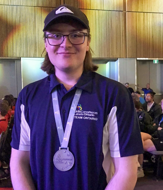 A man stands in an event conference room wearing a silver medal around his neck and smiling at the camera. 