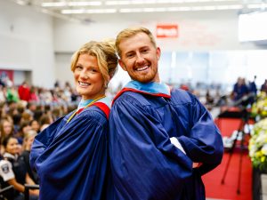 Two Brock University graduates stand shoulder-to-shoulder in front of a crowd of people.