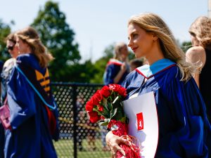 A young woman in a blue academic robe stands outside holding a bouquet of flowers and a diploma after a university graduation ceremony.