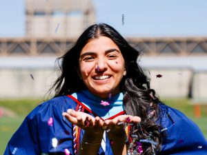 A close-up view of a young woman in a blue academic gown blowing a handful confetti into the camera outside after a university graduation ceremony.