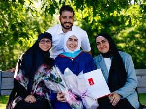 A young woman in a blue academic robe and white hijab poses with three loved ones outside after a university graduation ceremony. She is holding flowers and her diploma.