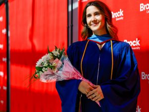 A young woman in a blue academic robe poses with a bouquet of flowers in front of a red background after a university graduation ceremony.