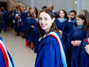 A young woman in a blue academic gown smiles for a photo in a sunny hallway. She is surrounded by other graduating students also wearing blue academic robes.