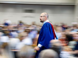 A man in a blue academic gown walks through the audience, which is out of focus, during a university graduation ceremony.