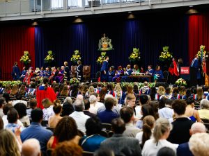 Brock University graduates walk across the stage while people sit on chairs behind them and a crowd of people in the audience look on.