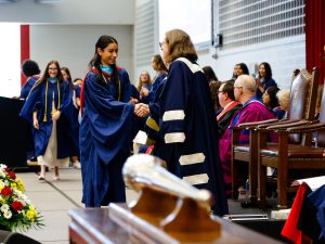 Brock University President Lesley Rigg shakes a graduate's hand on a stage while others seated in chairs look on.