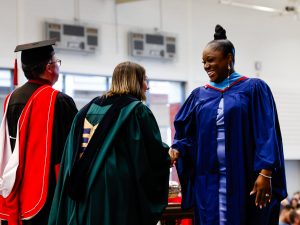 A female Brock University graduate shakes hands with a woman in a green gown who is standing beside a man in a red and black gown.