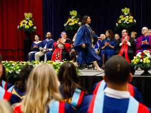 A female Brock University graduate walks across the stage in front of others sitting on chairs and a crowd of people in the audience.