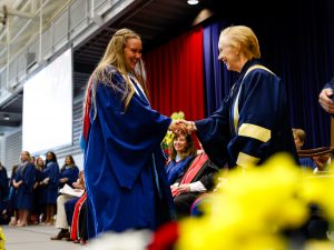 Two women shake hands on the stage during a convocation ceremony while others on the stage look on.