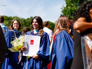 A young woman in a blue academic robe smiles for a photo while holding a diploma and bouquet of flowers outside a university graduation ceremony. She is surrounded by other graduates in gowns and their loved ones.