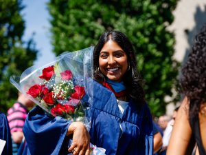 A smiling young woman in a blue academic robe poses with a bouquet of flowers outside after a university graduation ceremony.