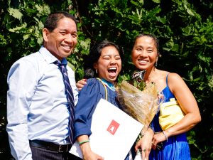 A smiling young woman holding a diploma and bouquet of flowers poses for a photo between two loved ones outside after a university graduation ceremony.