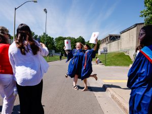 Two young women in blue academic robes smile and hold up their diplomas while loved ones take pictures outside after a university graduation ceremony.