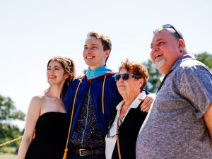 A young man in a blue academic gown poses for a photo with three smiling loved ones outside after a university graduation ceremony.