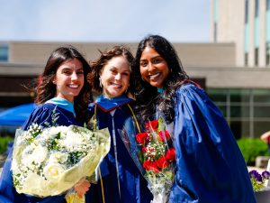 Three young in blue academic robes pose for a photo outside after a university graduation ceremony. Two of them are holding bouquets of flowers.