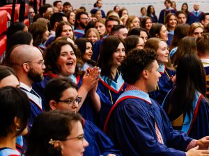 A crowd of graduates in graduation gowns gather in a gymnasium.