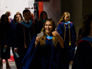 A woman in a graduation gown gives a thumbs up while walking in a hallway surrounded by other graduates.