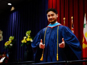 A man in a graduation gown raises his hands after crossing the Convocation stage.