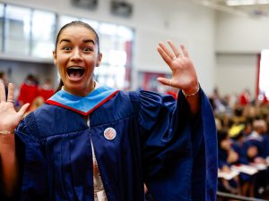 A woman in a graduation gown raises her hands in excitement while standing in a gymnasium full of graduates.