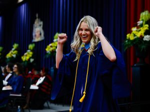 A woman in a graduation gown cheers with her arms in the air after walking across the Convocation stage.