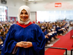 A woman wearing a graduation gown stands in front of a gymnasium of graduates who are seated watching a ceremony.