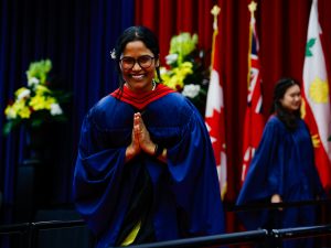 A woman wearing a graduation gown holds her hands together in a 'namaste' gesture.