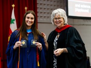 A woman in a graduation gown holds two gold medals in her hands while standing with a faculty member.