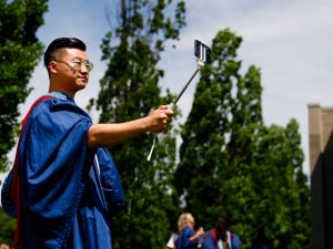 A man wearing a graduation gown holds a selfie stick and takes a photo of himself with a phone.