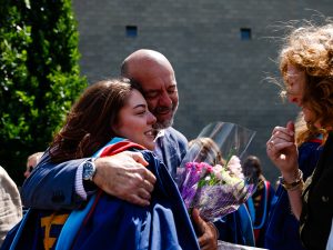 A man hugs a woman who is wearing a graduation gown and holding a bouquet of flowers.