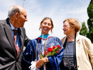 An older man and woman flank a young woman graduate who is wearing a graduation gown and holding a bouquet of flowers.