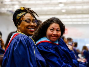 Two women in graduation gowns sit in a gymnasium with other grads.