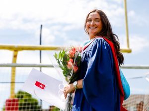 A woman in a graduation gown stands outside while holding a bouquet of flowers and a folder with her degree.