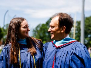 A woman and a man, each with graduation gowns on, look at each other while standing outside.