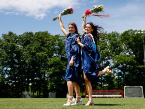 Two women in graduation gowns each raise a bouquet of flowers in the air with one arm while standing on a sports field.