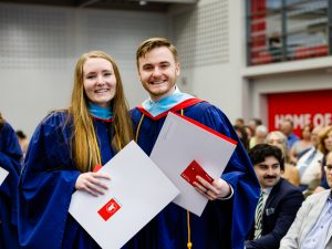 A woman and man, each in graduation gowns, holds folders containing their respective degrees.