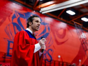 A man wearing academic regalia stands speaking into a microphone at Brock University’s Bob Davis Gymnasium.