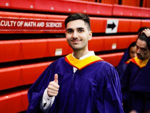 A man wearing a graduation gown gives a thumbs up in front of red bleachers.