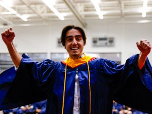 A man in a blue graduation gown celebrates by throwing his arms up while smiling during a university graduation ceremony.