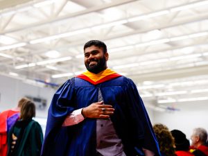 A man wearing a blue convocation gown smiles while walking across the graduation stage at Brock University.