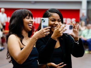 Two women wearing black hold up phones while laughing inside Ian Beddis Gymnasium at Brock University during a graduation ceremony.