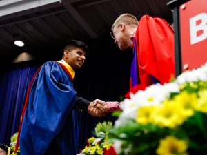 A man wearing a blue convocation gown shakes another man’s hand who is wearing a red convocation gown on stage at Brock University during a graduation ceremony.