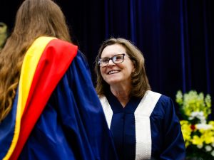 A woman wearing a blue and white ceremonial convocation gown smiles and shakes a woman’s hand during a university graduation ceremony.